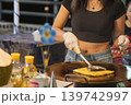 In Bangkok street market, a woman cooks roti on a hot surface while wearing gloves. She flips the dough with a spatula. The setting is lively with nearby vendors and customers. High quality photo 139742997