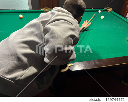 A boy bends over a billiards table, aiming his cue stick at the balls scattered on the green felt surface in a game room at night A boy bends over a billiards table, aiming his cue stick at the balls scattered on the green felt surface in a game room at night 139744371
