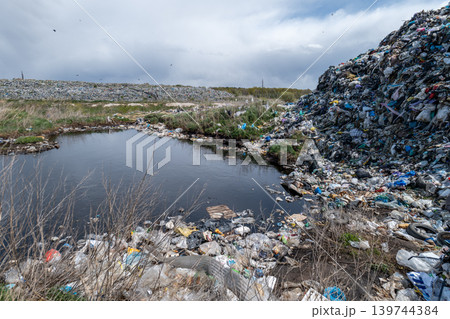 Waste and rubbish cover the ground near a small water body at a landfill site. Litter spreads across the area, creating an unpleasant scene Waste and rubbish cover the ground near a small water body at a landfill site. Litter spreads across the area, creating an unpleasant scene 139744384