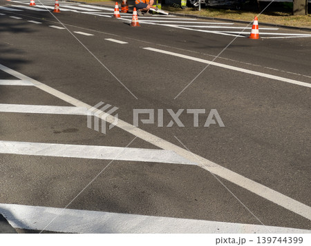 Workers are painting white road markings on a street. Safety cones are placed to guide traffic during the work Workers are painting white road markings on a street. Safety cones are placed to guide traffic during the work 139744399