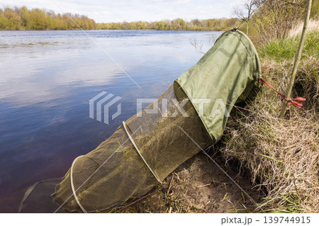 A green fishing net is set up by the riverbank. The net is partially submerged in the water during midday. The area has grass and trees nearby A green fishing net is set up by the riverbank. The net is partially submerged in the water during midday. The area has grass and trees nearby 139744915