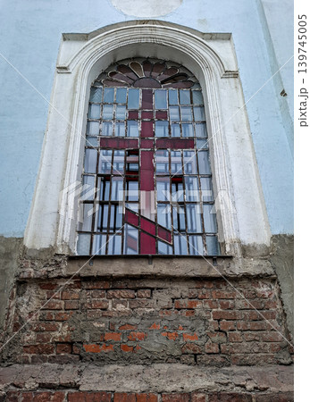 The arched window shows a large red Orthodox cross made of stained glass. It is set in a weathered brick wall of an ancient building. The colors are faded and the wall is cracked. The arched window shows a large red Orthodox cross made of stained glass. It is set in a weathered brick wall of an ancient building. The colors are faded and the wall is cracked. 139745005