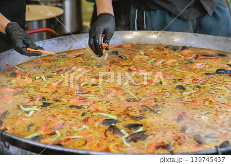 Seafood paella cooks in large pan with shrimp, mussels, broth, vegetables, oil, steam, gloved hands of chef. Concept of seafood paella for catering, menu, festival food, event dining, food preparation 139745437