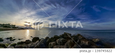 A wide scene shows a sunset over the sea. The sun sets on the horizon and reflects on the water. A rocky breakwater is in the foreground. Clouds fill the sky with interesting shapes. 139745822