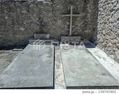 Two graves made of concrete sit side by side in an old cemetery. Stone and iron crosses mark the resting places under bright sunlight. The ground is bare and simple. Two graves made of concrete sit side by side in an old cemetery. Stone and iron crosses mark the resting places under bright sunlight. The ground is bare and simple. 139745902