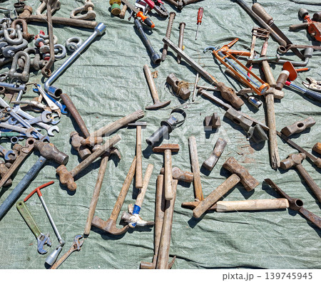 Various rusty hand tools are scattered on a wrinkled green tarp at an outdoor flea market. Old hammers, wrenches, screwdrivers, and pliers are visible among the items. 139745945