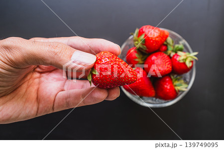 Hand holding glass bowl of fresh ripe red strawberries on dark black background Hand holding glass bowl of fresh ripe red strawberries on dark black background 139749095