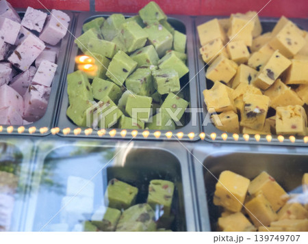 Colorful assortment of traditional Indian sweets displayed in a glass case, featuring pink, green, and yellow varieties with distinct shapes and textures 139749707