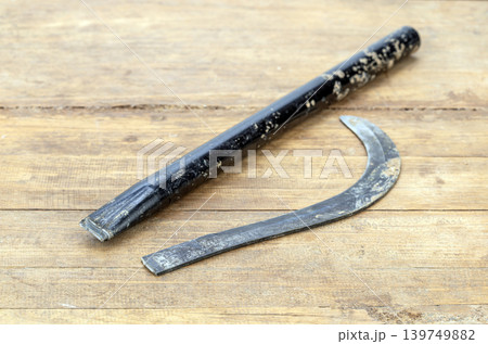 Old Worn Broken Iron Sickle Blade Separated from Its Black Handle Lying on a Rustic Wooden Surface, Showing Clear Break Point and Heavy Use Old Worn Broken Iron Sickle Blade Separated from Its Black Handle Lying on a Rustic Wooden Surface, Showing Clear Break Point and Heavy Use 139749882