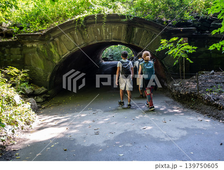 New york, usa august 18,2024. Three people walking through a historic stone arch tunnel, exploring the trails and vibrant greenery of central park in manhattan, new york New york, usa august 18,2024. Three people walking through a historic stone arch tunnel, exploring the trails and vibrant greenery of central park in manhattan, new york 139750605