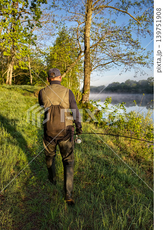 A person walks along the riverbank in the morning light, holding a fishing rod. Trees surround the area, and a gentle mist rises off the water 139751908