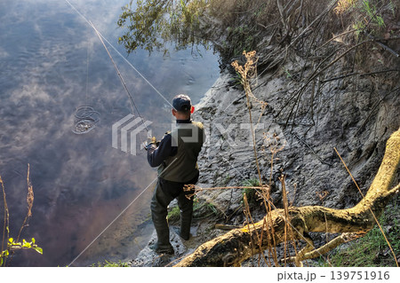 A man stands on the riverbank holding a fishing rod. He is focused on the water while trying to catch fish. It is a clear day with some clouds A man stands on the riverbank holding a fishing rod. He is focused on the water while trying to catch fish. It is a clear day with some clouds 139751916