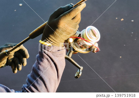 A person grips a fishing rod with a reel in hand by the river as the sun rises. The dark water reflects the light of dawn creating a still scene 139751918