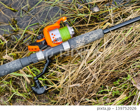 A fishing rod and reel rest on dry grass by a body of water. The bright reel contrasts with the earth tones around it. This scene shows a moment before fishing starts A fishing rod and reel rest on dry grass by a body of water. The bright reel contrasts with the earth tones around it. This scene shows a moment before fishing starts 139752026