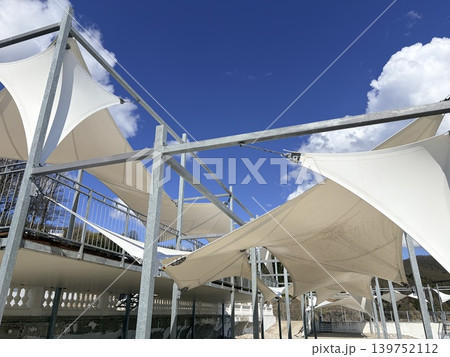 a modern outdoor shading structure featuring multiple white tensile fabric canopies stretched taut over a framework of galvanized steel beams and vertical posts. a modern outdoor shading structure featuring multiple white tensile fabric canopies stretched taut over a framework of galvanized steel beams and vertical posts. 139752112