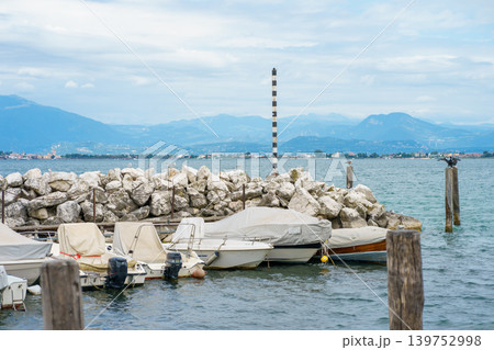 Small motorboats covered and docked at calm pier on Lake Garda, Italy, with scenic mountains and cloudy sky creating peaceful waterfront atmosphere Small motorboats covered and docked at calm pier on Lake Garda, Italy, with scenic mountains and cloudy sky creating peaceful waterfront atmosphere 139752998