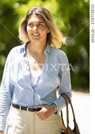 Woman walks with confidence in a garden during daytime while holding a bag and dressed in light colors Woman walks with confidence in a garden during daytime while holding a bag and dressed in light colors 139753610