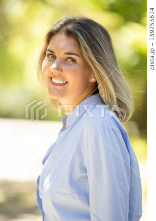 Young woman smiles while standing outdoors in a green space during daytime 139753614