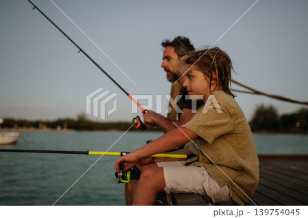 Father and son fishing together from wooden pier at sunset during family leisure time. Father and son fishing together from wooden pier at sunset during family leisure time. 139754045