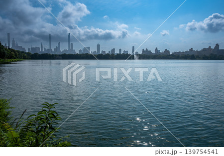 New york, usa august 18,2024. Central park's jacqueline kennedy onassis reservoir reflecting manhattan skyline under a cloudy blue sky, serene urban water view 139754541