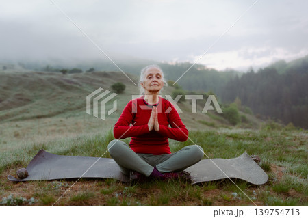 Senior woman meditating in nature on early morning with fog and mountains in background. Senior woman meditating in nature on early morning with fog and mountains in background. 139754713