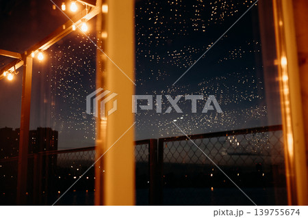 Cozy night rooftop atmosphere with hanging string lights and starry sky reflections. Warm glowing light bulbs against dark city skyline background. Magical evening terrace vibe and festive mood. 139755674
