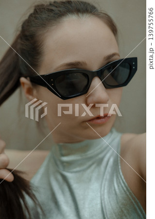 Young woman in stylish silver metallic top and trendy cat-eye sunglasses posing outdoors. Authentic film grain aesthetic, Gen Z fashion style, retro-futuristic urban look with neutral background. 139755766