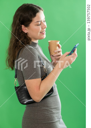Young woman scrolling smartphone and drinking coffee in front of green screen Young woman scrolling smartphone and drinking coffee in front of green screen 139757502