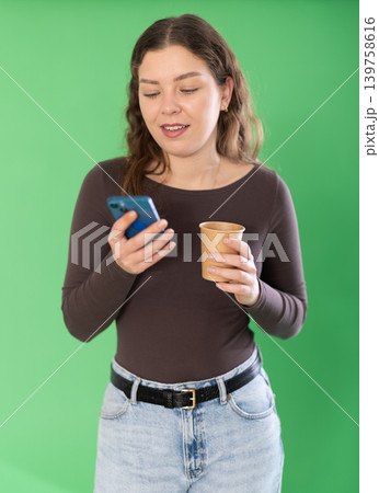 Young woman scrolling smartphone and drinking coffee in front of green screen Young woman scrolling smartphone and drinking coffee in front of green screen 139758616