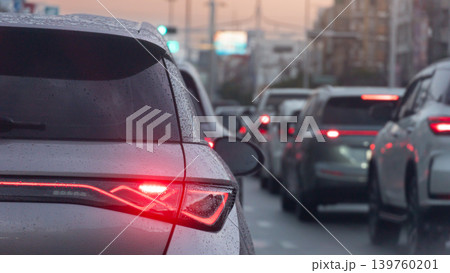 Behind a white car with its red brake lights illuminated during evening traffic with drop rain on car. Vehicles stretches ahead on a crowded city street as dusk begins to settle urban landscape. 139760201