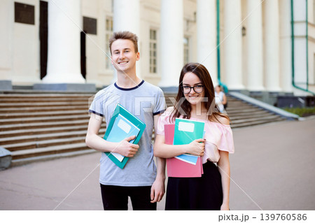 Two exemplary students in front of old beautiful conventional university with high columns posing with learning materials dressed in casual clothes looks happy and encouraged to study Two exemplary students in front of old beautiful conventional university with high columns posing with learning materials dressed in casual clothes looks happy and encouraged to study 139760586