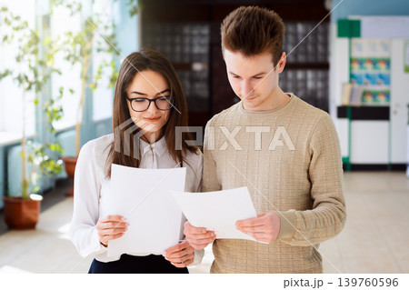 Portrait of students with sheets of paper with results of their tests. Male and female learners standing in corridor of university with pleased expression on their face looking down to results of work 139760596