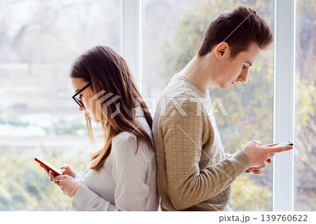 Couple of young students sitting next to one another and using mobile phones having no expression, concerned to their phones, have no interest in one another, problem of constant using modern gadgets 139760622