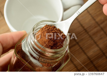 Man`s hand taking some ground coffee from coffee jar. Empty cup stands on the brown background. Making coffee Man`s hand taking some ground coffee from coffee jar. Empty cup stands on the brown background. Making coffee 139760647