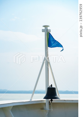 Blue flag on boat mast with metal bell against soft sky background, minimalistic nautical composition with calm lake horizon and light tones Blue flag on boat mast with metal bell against soft sky background, minimalistic nautical composition with calm lake horizon and light tones 139762959