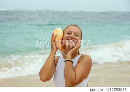 Woman smiles while holding a seashell at the beach Woman smiles while holding a seashell at the beach 139763085