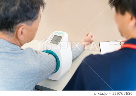血圧計を使い血圧測定をするシニア男性と記録をとる看護師,介護士の男性スタッフ 血圧計を使い血圧測定をするシニア男性と記録をとる看護師,介護士の男性スタッフ 139763656