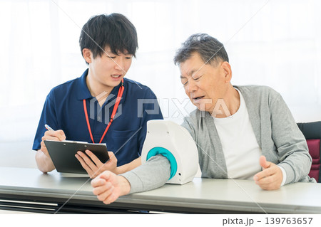 血圧計を使い血圧測定をするシニア男性と記録をとる看護師,介護士の男性スタッフ 血圧計を使い血圧測定をするシニア男性と記録をとる看護師,介護士の男性スタッフ 139763657