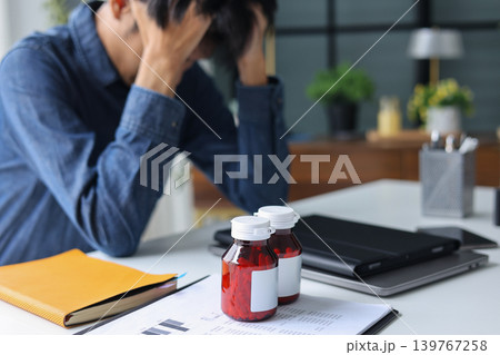Male employee suffering from migraine and stress in office with medicine bottles on the table. 139767258