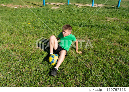 Boy rests on the grass, holding a soccer ball. A sunny day outdoors. Boy rests on the grass, holding a soccer ball. A sunny day outdoors. 139767538