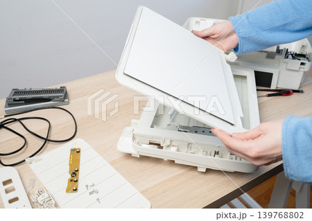 Hands carefully disassembling a flatbed scanner on a wooden desk, showing internal parts, cables and tools during repair and maintenance in a home office or small workplace setting. Hands carefully disassembling a flatbed scanner on a wooden desk, showing internal parts, cables and tools during repair and maintenance in a home office or small workplace setting. 139768802