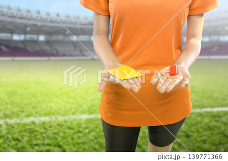 A referee in an orange shirt stands on a football pitch holding a yellow penalty card and a red whistle, ready to officiate a match in a large stadium A referee in an orange shirt stands on a football pitch holding a yellow penalty card and a red whistle, ready to officiate a match in a large stadium 139771366
