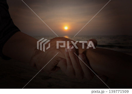 A close-up shot showing two hands clasped in a supportive gesture against the backdrop of a beautiful sunset over the calm ocean waves at dusk A close-up shot showing two hands clasped in a supportive gesture against the backdrop of a beautiful sunset over the calm ocean waves at dusk 139771509