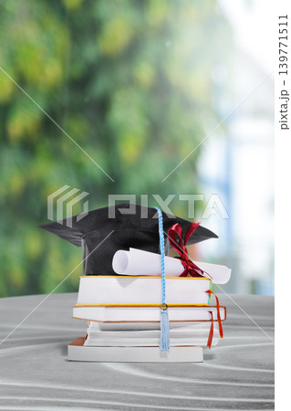 A black graduation mortarboard with a blue tassel sits atop a stack of books and a diploma tied with a red ribbon, set against a blurred green background A black graduation mortarboard with a blue tassel sits atop a stack of books and a diploma tied with a red ribbon, set against a blurred green background 139771511