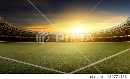 A wide-angle view of a professional football stadium at sunset. The empty arena features a lush green field under a dramatic golden hour sky with a bright lens flare 139771538