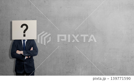 A businessman in a suit stands with arms crossed against a gray concrete wall, his head obscured by a sign featuring a large black question mark, symbolizing anonymity, doubt, or curiosity A businessman in a suit stands with arms crossed against a gray concrete wall, his head obscured by a sign featuring a large black question mark, symbolizing anonymity, doubt, or curiosity 139771590