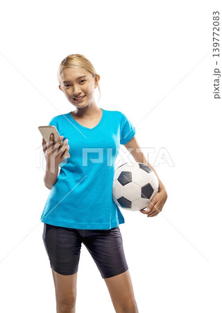 A happy young Indonesian southeast asian woman in a blue t-shirt holding a football and checking her mobile phone, isolated on a white background. Ideal for sports betting or app concepts 139772083