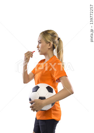 A young Indonesian southeast asian woman in an orange jersey and black shorts holds a soccer ball and blows a whistle against a white background A young Indonesian southeast asian woman in an orange jersey and black shorts holds a soccer ball and blows a whistle against a white background 139772257