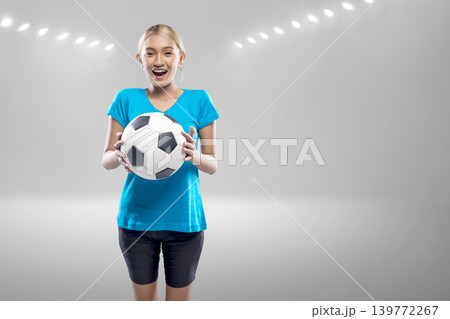 A joyful young Indonesian southeast asian woman with a bright smile holds a soccer ball against a blurred stadium background with glowing lights. Perfect for sports and fitness themes A joyful young Indonesian southeast asian woman with a bright smile holds a soccer ball against a blurred stadium background with glowing lights. Perfect for sports and fitness themes 139772267