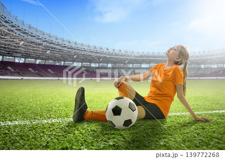 Young Indonesian southeast asian woman soccer athlete resting on a green pitch in a large empty stadium after a training session or game, showing fatigue and emotion with a football beside her Young Indonesian southeast asian woman soccer athlete resting on a green pitch in a large empty stadium after a training session or game, showing fatigue and emotion with a football beside her 139772268
